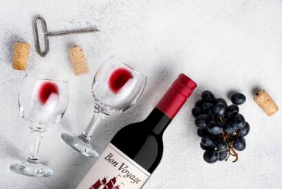 Bon Voyage red wine bottle with wine glasses, a corkscrew, corks and black grapes on a white surface