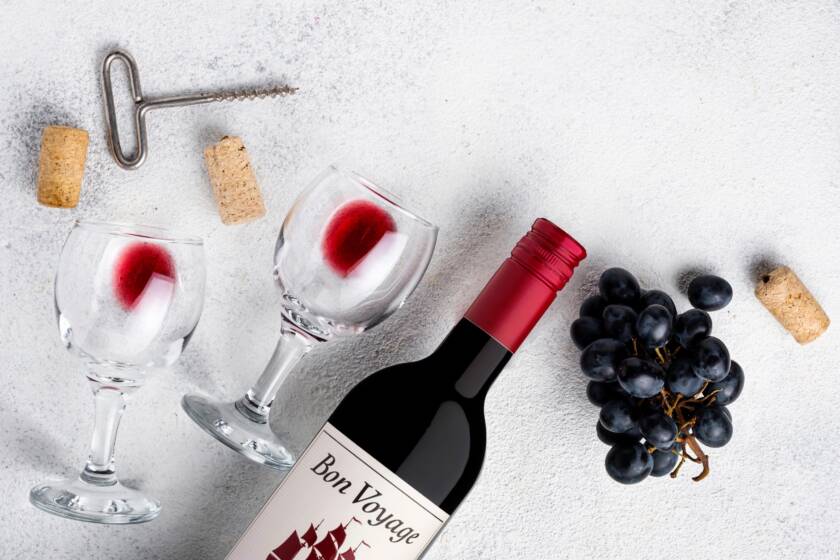 Bon Voyage red wine bottle with wine glasses, a corkscrew, corks and black grapes on a white surface