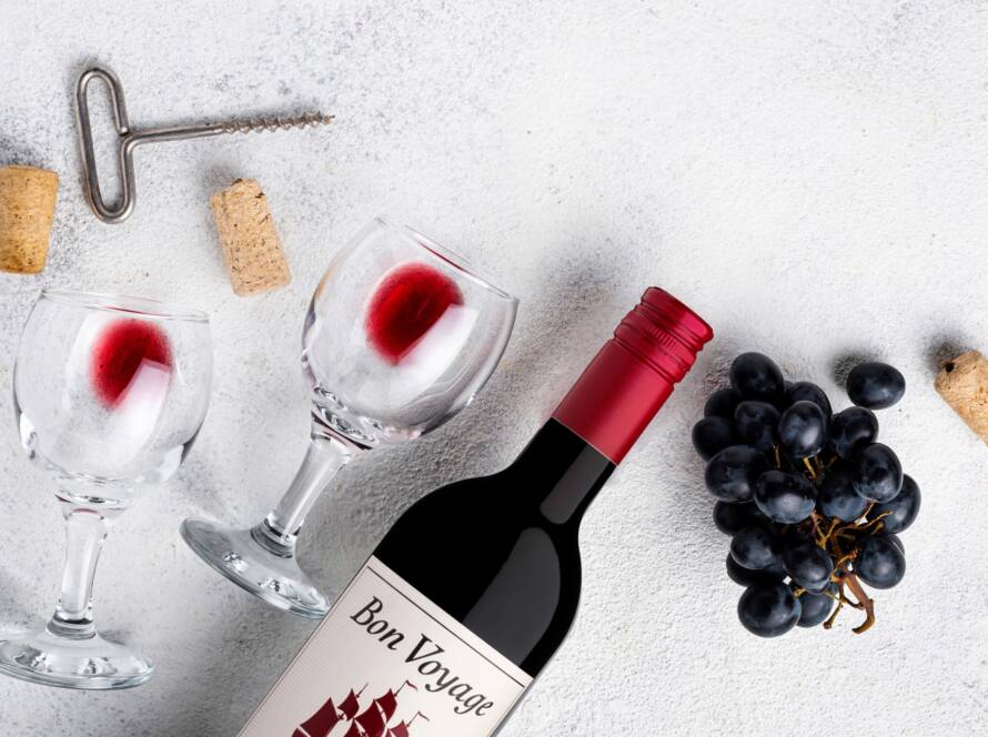 Bon Voyage red wine bottle with wine glasses, a corkscrew, corks and black grapes on a white surface